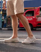 Person wearing light-colored slip-on shoes and beige shorts on a street with a red truck in the background.